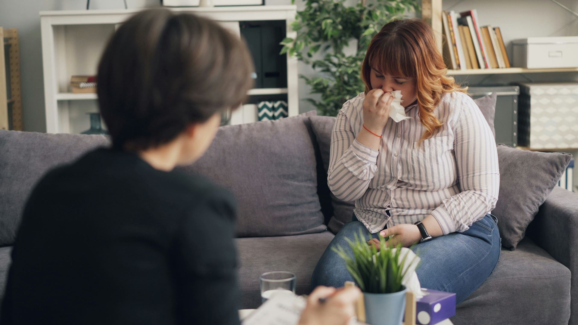 a woman sat on a sofa sneezing into a tissue with another woman watching