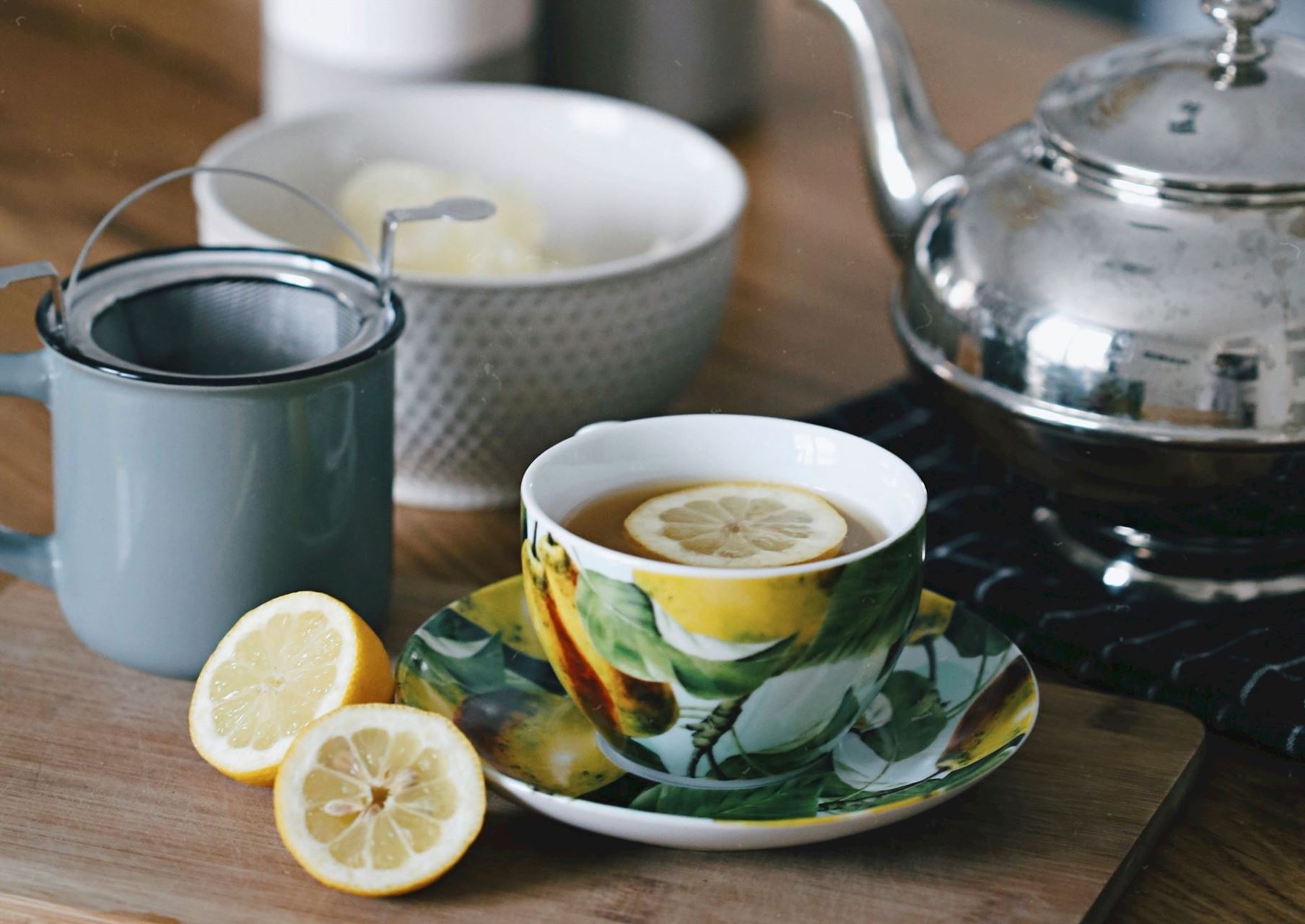 a cup of tea in a china cup with a slice lemon in it, two halves of lemons on the side, a sugar bowl and a silver teapot
